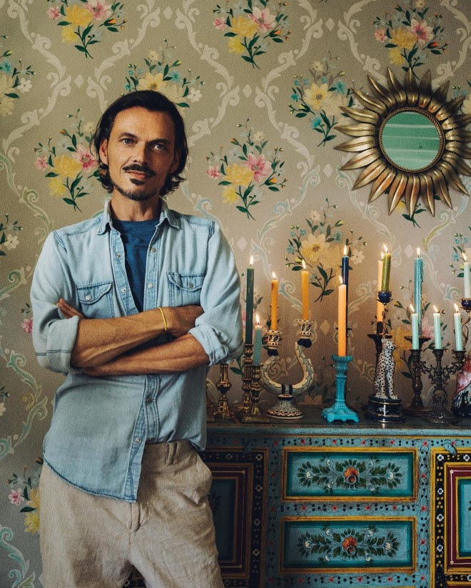 A picture of designer Matthew Williamson, standing next to a blue dresser with a wide array of candles and candlesticks on the top. Behind him in a ornate floral wallpaper with a sunburst mirror